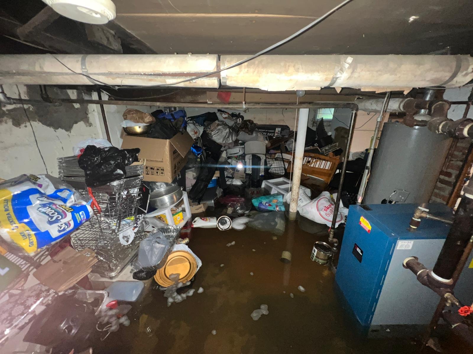 Anajur flood response, same event showing a basement utility area with a blue residential boiler and adjacent water heater partially submerged in turbid floodwater, overhead pipes and structural columns visible, storage items displaced throughout the space