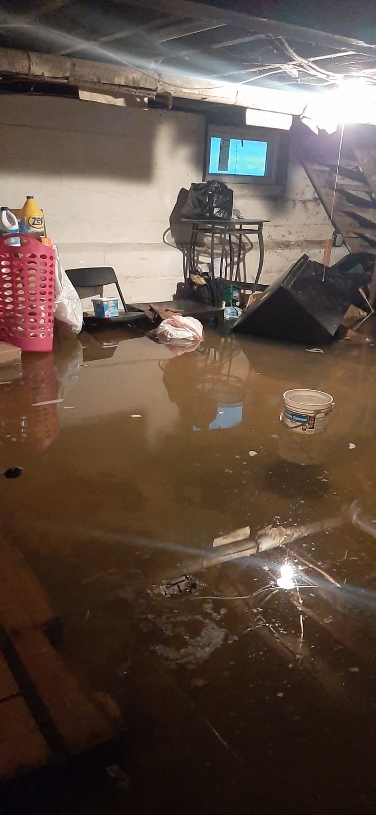 Anajur flood response, same event from a different angle, daylight entering through a cellar window above the water line, household items including a laundry basket and chair displaced and partially submerged by rising water